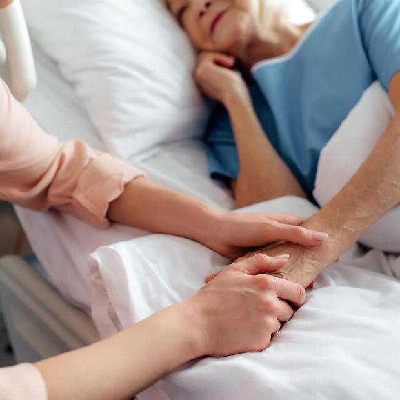 Senior woman resting in bed holding hands with young female carer
